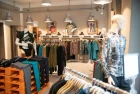 Modern salesroom by Chapati Design with sustainable menswear. Wooden shelves and clothes rails display colorful shirts, jackets and pants. Industrial lamps provide atmospheric lighting. Mannequins in stylish clothing and decorative pictures on the wall can be seen in the background.