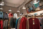 A clothes rack takes center stage, stocked with sustainable shirts and jackets in warm colors. Other items of clothing hang on the walls, including hoodies and tops in shades of blue and red. A large mirror reflects the ceiling with modern industrial lamps and enhances the spatial effect.